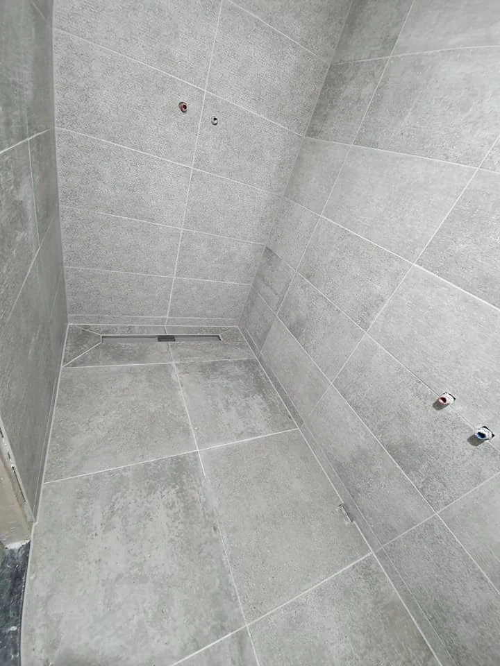 Grijs tegelwerk in een douche met vloerafvoer en aansluitpunten voor sanitair.
